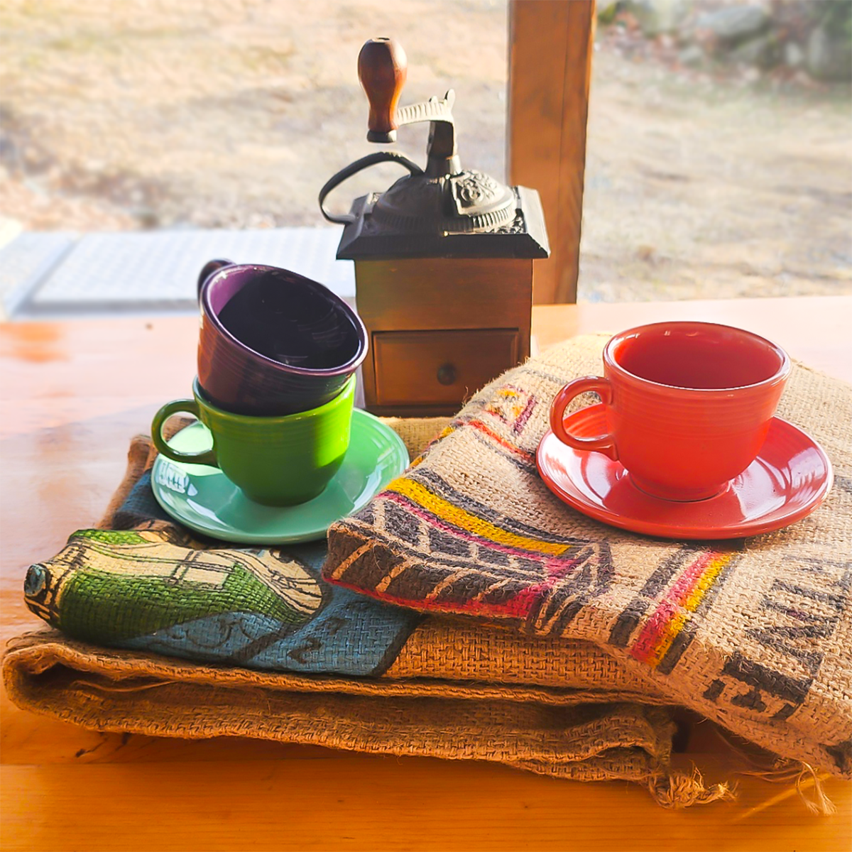 A set of colorful coffee cups and saucers displayed on top of a jute coffee bag with a wooden coffee grinder in the background.