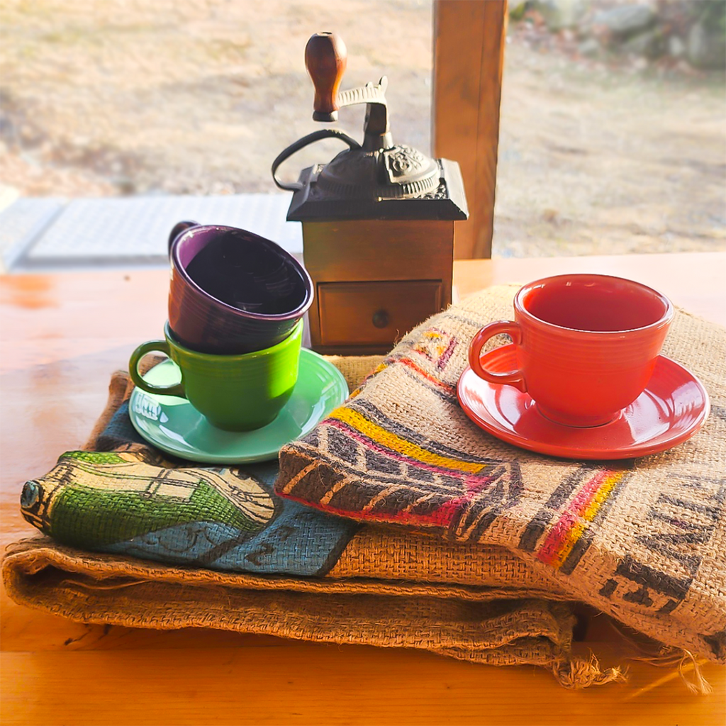 A set of colorful coffee cups and saucers displayed on top of a jute coffee bag with a wooden coffee grinder in the background.